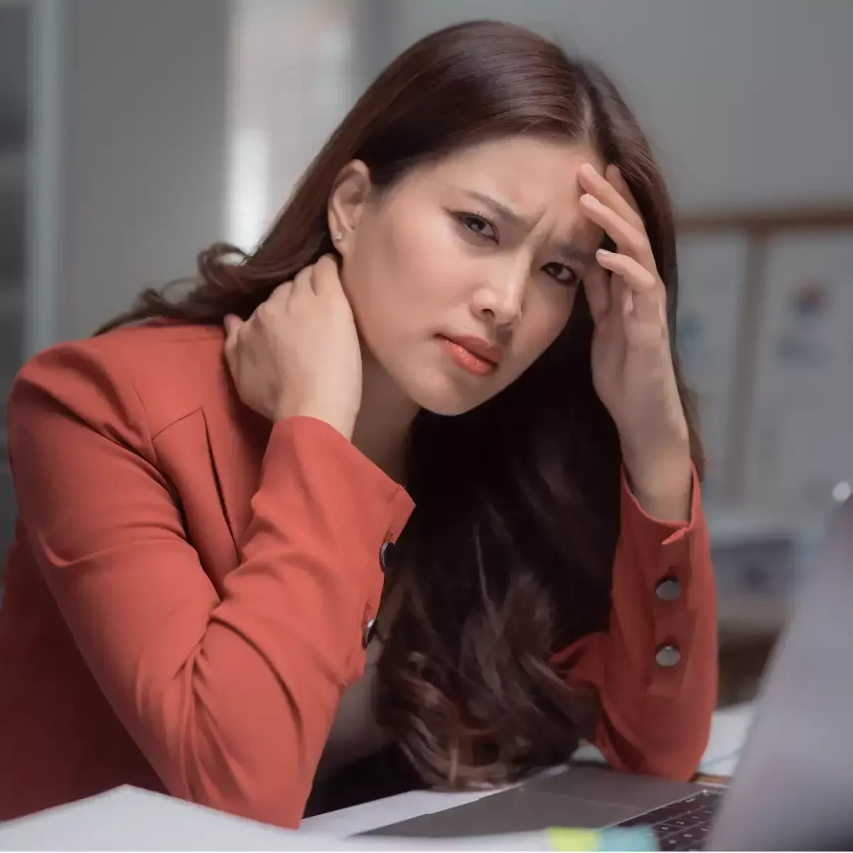 Office worker holding her neck and forehead while experiencing neck pain and headache at her desk.